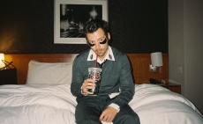 Harry Styles sits on a bed, sipping a drink with dark, decorative patches beneath his eyes