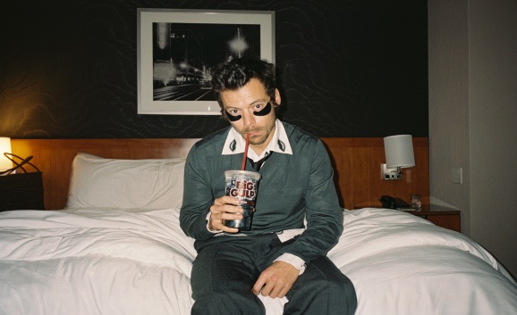 Harry Styles sits on a bed, sipping a drink with dark, decorative patches beneath his eyes