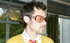 Harry Styles wears a yellow jumper with a salmon pink tie and transparent orange sunglasses