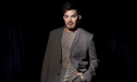 Adam Lambert wears a grey blazer and poses in a press shot